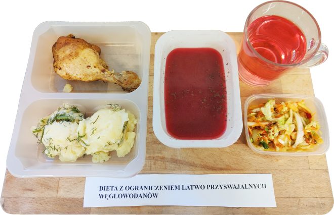 Zestaw obiadowy na tacy: w pojemniku znajduje się pieczony podudzie z kurczaka oraz purée ziemniaczane posypane koperkiem. Obok zupa – barszcz czerwony oraz kubek z napojem owocowym o czerwonej barwie. W osobnym pojemniczku surówka z kapusty pekińskiej, marchwi i papryki. Przed tacką widoczny napis „DIETA Z OGRANICZENIEM ŁATWO PRZYSWAJALNYCH WĘGLOWODANÓW”