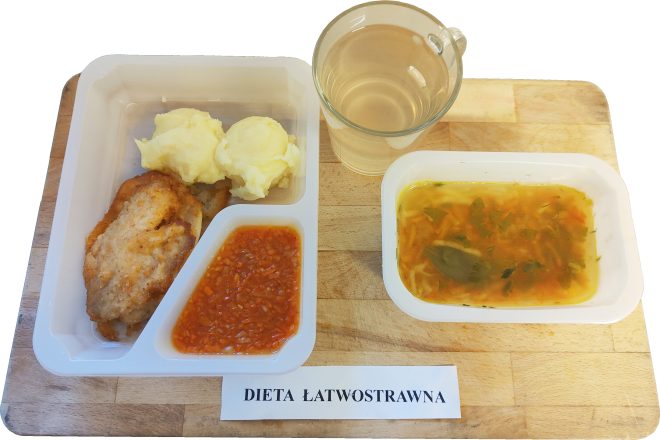 Na tacy ułożony zestaw obiadowy w pojemniku jednorazowym. W głównej części widać panierowany filet z kurczaka z porcją purée ziemniaczanego oraz sosem pomidorowym. Obok pojemnik z lekkim bulionem warzywnym z makaronem i natką pietruszki. Do zestawu dołączony kubek z napojem ziołowym lub herbatą. Na kartce przed tacką napis „DIETA ŁATWOSTRAWNA”
