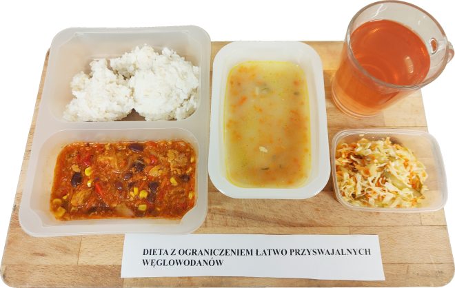 Na tacy znajduje się zestaw obiadowy w pojemnikach jednorazowych. W skład posiłku wchodzi zupa jarzynowa, ryż biały oraz danie główne z mięsem drobiowym, czerwoną fasolą, kukurydzą i papryką w sosie pomidorowym. Obok pojemników ustawiono surówkę z białej kapusty i marchwi oraz szklankę kompotu o różowym zabarwieniu. Całość oznaczona jest etykietą z napisem: „Dieta z ograniczeniem łatwo przyswajalnych węglowodanów