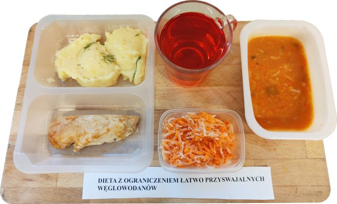 Zestaw obiadowy w pudełkach dla diety z ograniczeniem łatwo przyswajalnych węglowodanów: porcja puree ziemniaczanego z koperkiem, gotowany kawałek piersi kurczaka, surówka z marchewki i białej rzodkwi, zupa jarzynowa oraz szklanka czerwonego napoju. Pod tacą widoczna etykieta z nazwą diety