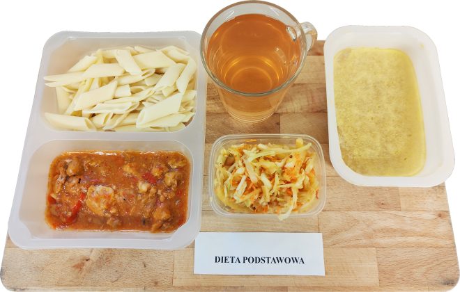 Tacka z posiłkiem szpitalnym w diecie podstawowej: gotowany makaron penne, duszone mięso w sosie z warzywami, surówka z kapusty i marchewki, kubek herbaty oraz miseczka z zupą o jasnym, kremowym kolorze. Na desce znajduje się kartka z napisem „Dieta podstawowa”