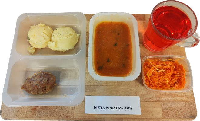 Na drewnianej desce stoi zestaw obiadowy w plastikowych pojemnikach opisany jako „Dieta podstawowa”. Widać porcję ziemniaków puree, kotlet mielony, pojemnik z zupą pomidorową, surówkę z marchwi oraz szklankę czerwonego kompotu