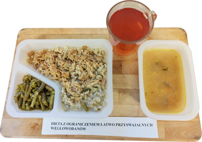 Na tacy ustawiono pojemnik z kaszą i drobno rozdrobnionym mięsem, porcję zielonej fasolki oraz zupę w oddzielnym pojemniku. Obok stoi szklanka czerwonego napoju. Pod posiłkiem znajduje się etykieta „Dieta z ograniczeniem łatwo przyswajalnych węglowodanów”