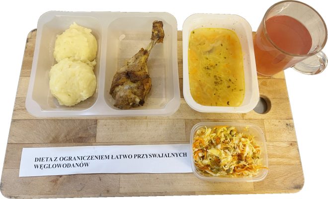 Zestaw obiadowy w diecie z ograniczeniem łatwo przyswajalnych węglowodanów. Na tacy znajdują się: porcja puree ziemniaczanego, pieczona ćwiartka kurczaka, miseczka z lekką zupą jarzynową, surówka z białej kapusty i marchwi oraz kubek czerwonego napoju owocowego. Przed tacą widoczny jest opis diety