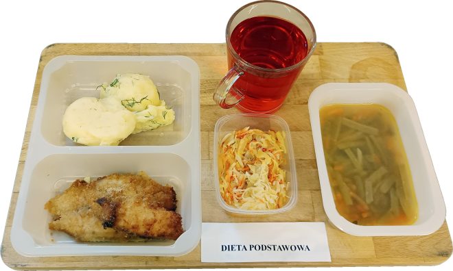 Na drewnianej tacy ustawiono zestaw obiadowy w plastikowych pojemnikach. W jednym z nich znajduje się porcja puree ziemniaczanego z koperkiem. W drugim leży panierowany kawałek mięsa. Obok stoi mały pojemnik z surówką z białej kapusty i marchewki oraz oddzielny pojemnik z zupą jarzynową. Za zestawem znajduje się szklanka czerwonego napoju. Przed posiłkiem widoczna jest etykieta z napisem „Dieta podstawowa”