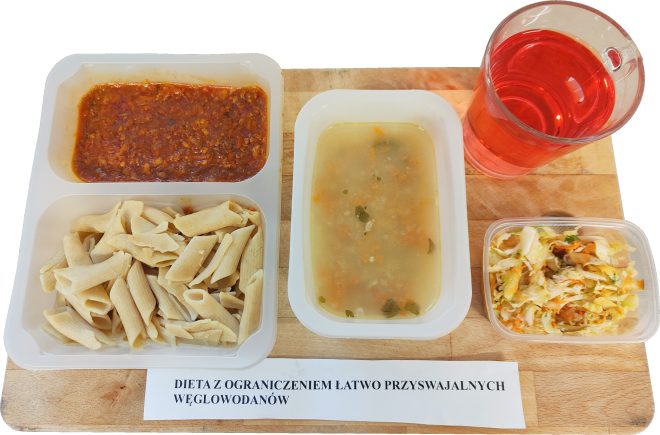 Obiad w diecie z ograniczeniem łatwo przyswajalnych węglowodanów: tacka z makaronem penne i sosem pomidorowo-warzywnym, pojemnik z lekką zupą jarzynową oraz sałatka z kapusty i marchwi. Obok stoi kubek czerwonego napoju. Na tacy znajduje się podpis «Dieta z ograniczeniem łatwo przyswajalnych węglowodanów