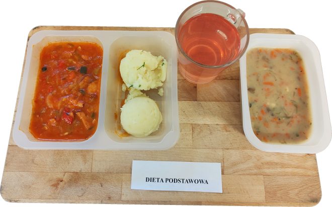 Zestaw obiadowy w diecie podstawowej podany w trzech plastikowych przegródkach na drewnianej tacy: gulasz warzywno-mięsny w czerwonym sosie, obok dwie porcje ziemniaków puree z zieleniną oraz miseczka jasnej zupy jarzynowej z marchewką i koperkiem. Do zestawu dołączono szklankę czerwonego kompotu