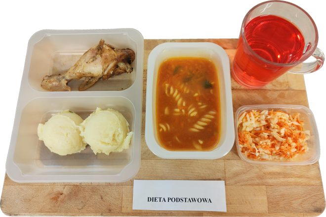 Zestaw obiadowy w diecie podstawowej: porcja zupy z makaronem w pojemniku, udko z kurczaka, dwie kulki puree ziemniaczanego, surówka z białej kapusty i marchwi oraz szklanka czerwonego napoju. Posiłki podane na desce z podpisem „Dieta podstawowa”