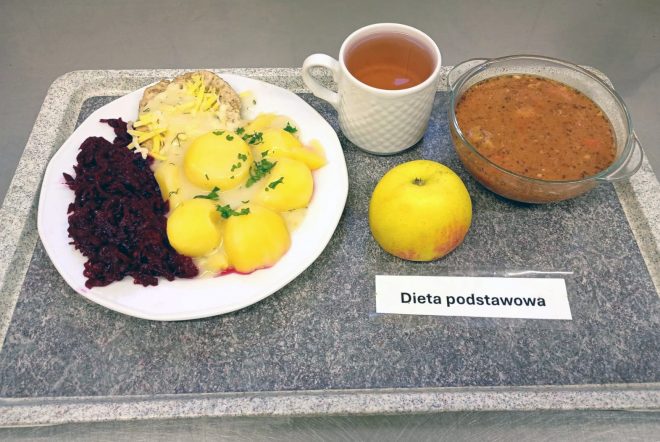 Na tacy widoczny jest zestaw obiadowy w diecie podstawowej. Na białym talerzu podano porcję ziemniaków z sosem i posypką z natki pietruszki, gotowane buraczki oraz kawałek mięsa z tartym serem. Obok znajduje się szklana miseczka z zupą jarzynową lub grochową, filiżanka z herbatą oraz jabłko. Całość ustawiona jest na szarej tacy, z karteczką z napisem „Dieta podstawowa