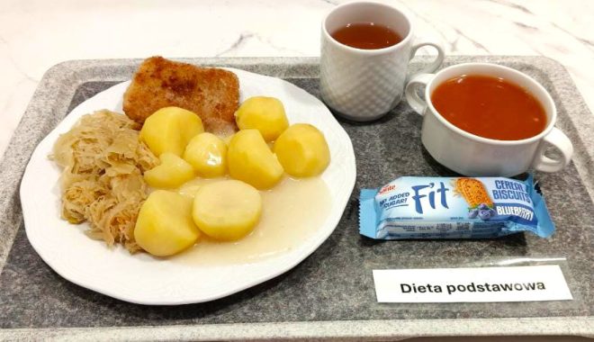 Na białym talerzu podane ziemniaki z sosem, surówka z duszonej kapusty oraz panierowany kawałek ryby. Obok na tacy filiżanka herbaty, miseczka z zupą pomidorową i batonik zbożowy „Fit”. Całość ustawiona na tacy z etykietą „Dieta podstawowa”