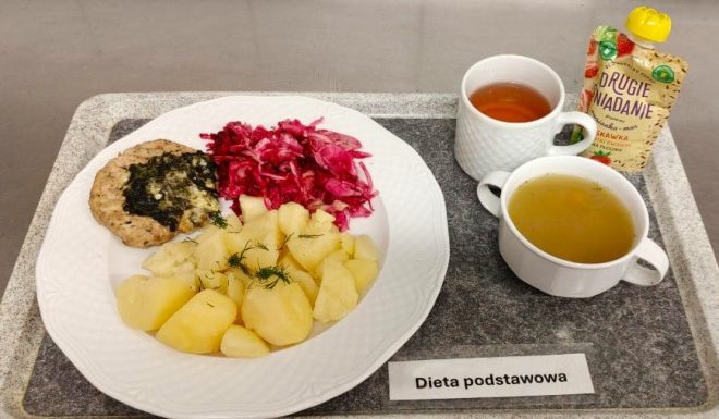 Na talerzu porcja gotowanych ziemniaków posypanych koperkiem, grillowany kotlet drobiowy ze szpinakiem, surówka z czerwonej kapusty i marchwi. Obok filiżanka z herbatą, miseczka z zupą oraz mus owocowy w saszetce. Etykieta na tacy: „Dieta podstawowa”