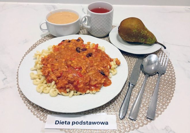 Talerz z obiadem w diecie podstawowej: makaron podany z gęstym sosem meksykańskim zawierającym warzywa i czerwoną fasolę. Obok stoi miseczka z zupą krem, kubek z herbatą oraz gruszka ułożona na małym talerzyku