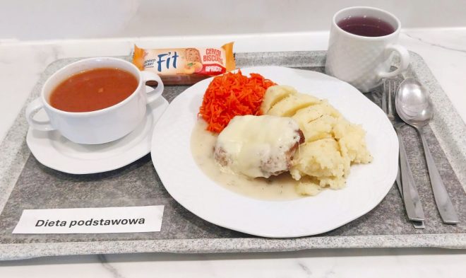 Zestaw obiadowy zawiera zupę pomidorową podaną w białej miseczce oraz talerz z puree ziemniaczanym, kotletem w białym sosie i porcją tartej marchewki. Za talerzem leży baton zbożowy, a obok znajduje się herbata. Całość ułożona jest na tacy z podpisem „Dieta podstawowa”
