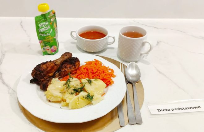 Zestaw obiadowy na diecie podstawowej: pieczone pałki z kurczaka, ziemniaki z koperkiem, surówka z marchewki, podane z czerwonym barszczem i herbatą; obok deser owocowy w tubce