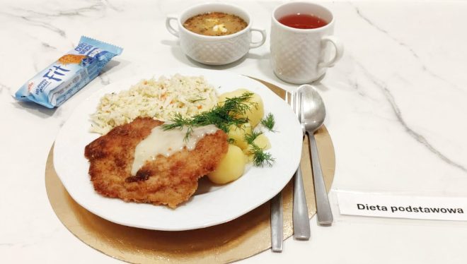 Talerz z panierowanym kotletem polanym jasnym sosem, gotowanymi ziemniakami z koperkiem i surówką z białej kapusty. Obok znajduje się baton zbożowy. W tle biała miseczka z zupą oraz kubek czerwonej herbaty. Po prawej stronie widoczna karteczka z napisem „Dieta podstawowa”