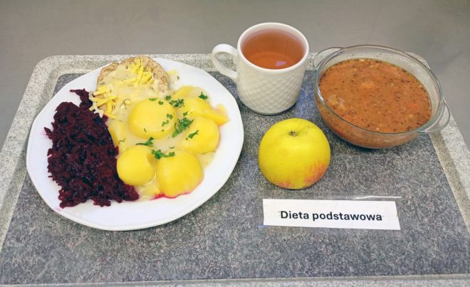 Na zdjęciu zestaw obiadowy w ramach diety podstawowej, podany na tacy. Na talerzu znajdują się gotowane ziemniaki z sosem, kotlet mielony posypany tartym serem oraz buraczki na ciepło. Obok położono jabłko, filiżankę z kompotem lub herbatą oraz miskę z zupą jarzynową. Przed zestawem widoczna jest kartka z napisem „Dieta podstawowa”