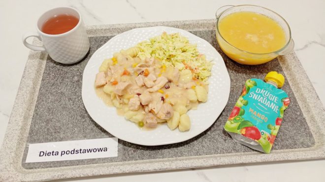 Na tacy oznaczonej etykietą „Dieta podstawowa” znajduje się talerz z kluseczkami gnocchi pod sosem z kawałkami kurczaka, warzywami i surówką z kapusty. Obok stoi miseczka z zupą jarzynową lub kremem oraz kubek z herbatą. Na tacy znajduje się także mus owocowy w saszetce o smaku mango i jabłka