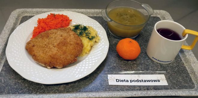 Na tacy z napisem Dieta podstawowa znajduje się talerz z panierowanym kotletem schabowym, porcją ziemniaków puree posypanych koperkiem oraz surówką z marchwi. Obok ustawiono miseczkę z klarowną zupą, kubek z herbatą owocową i mandarynkę
