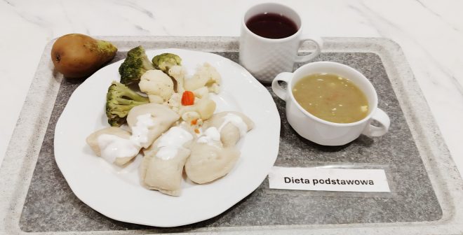 Na tacy z napisem „Dieta podstawowa” znajduje się zestaw obiadowy. Na głównym talerzu podano pierogi polane śmietaną oraz porcję gotowanych warzyw – brokułów, kalafiora i marchewki. Obok stoi filiżanka z zupą, druga filiżanka z kompotem oraz gruszka jako dodatek owocowy