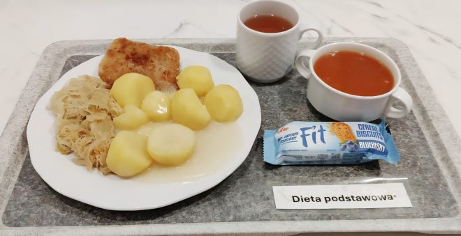 Na tacy z napisem „Dieta podstawowa” podano obiad: na dużym talerzu znajdują się ziemniaki z sosem, duszona kapusta i kotlet smażony. Obok stoją dwa kubki – jeden z herbatą, drugi z zupą pomidorową – oraz zapakowany batonik owsiany „Fit” z borówką. Całość ułożona na szarej tacy na tle białego blatu