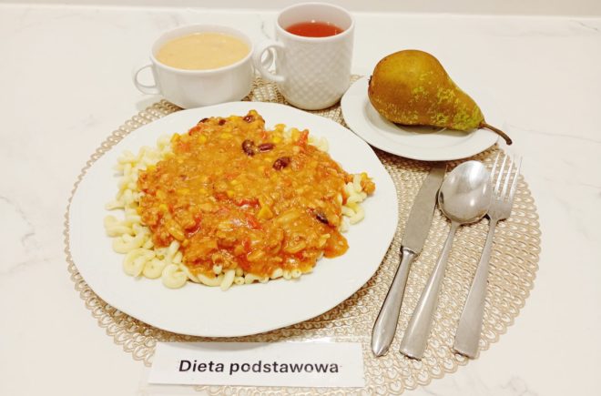 Na białym talerzu makaron rurki z sosem bolońskim z mięsem mielonym, fasolą, kukurydzą i pomidorami. Obok miseczka z zupą kremem, filiżanka z herbatą i talerzyk z gruszką. Zestaw ułożony na tacy z napisem „Dieta podstawowa”