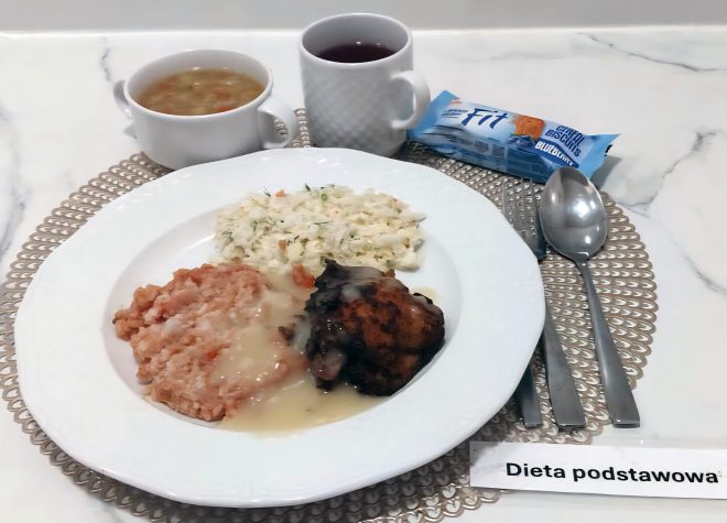 Talerz z obiadem w diecie podstawowej: porcja mięsa w jasnym sosie, ryż wymieszany z warzywami oraz surówka z białej kapusty z koperkiem. Obok znajduje się miseczka z zupą jarzynową, kubek z herbatą oraz pakowany herbatnik. Zestaw ułożony na jasnym, dekoracyjnym podkładzie