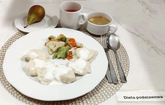 Zestaw obiadowy oznaczony jako „Dieta podstawowa”, podany na białej zastawie. Na talerzu znajdują się pierogi polane śmietaną oraz porcja gotowanych warzyw: brokułów, kalafiora i marchwi. Obok stoją dwie filiżanki — jedna z czerwonym napojem, druga z jasną zupą. W tle widoczna jest także gruszka na małym talerzyku. Na stole leżą sztućce