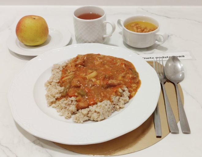 Zestaw obiadowy w diecie podstawowej. Na dużym białym talerzu znajduje się porcja kaszy jęczmiennej polanej gulaszem warzywno-mięsnym w sosie pomidorowym. W tle stoją: kubek herbaty w białej porcelanowej filiżance, miseczka z zupą jarzynową oraz talerzyk z jabłkiem. Po prawej stronie ułożone są sztućce na złotej podkładce. Pod talerzem widoczna etykieta z napisem ‘Dieta podstawowa’