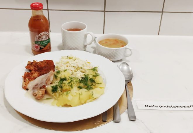 Talerz z pieczoną piersią z kurczaka, gotowanymi ziemniakami posypanymi koperkiem oraz surówką z białej kapusty. Obok filiżanka herbaty, miseczka z zupą jarzynową i butelka soku marchwiowo-jabłkowego. Widoczny napis ‘Dieta podstawowa’