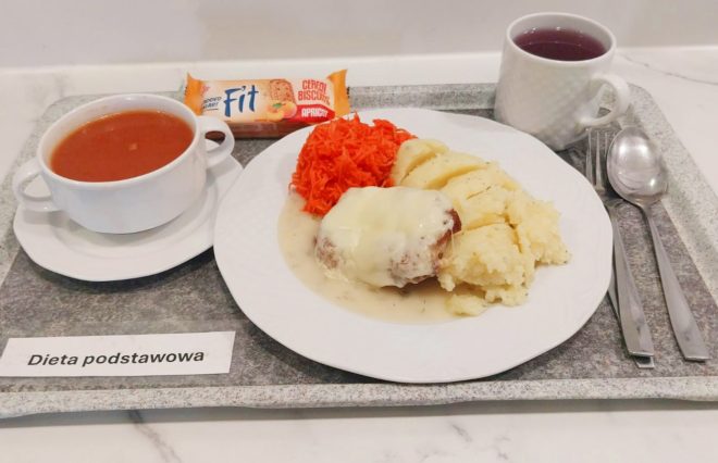 Na tacy znajduje się talerz z obiadem: kotlet drobiowy zapiekany z serem w sosie, ziemniaki puree oraz surówka z tartych marchewek. Obok stoi miseczka z zupą pomidorową oraz kubek z herbatą. W zestawie znajduje się także baton zbożowy. Przy tacy leżą sztućce, a całość opisana jest etykietą „Dieta podstawowa”
