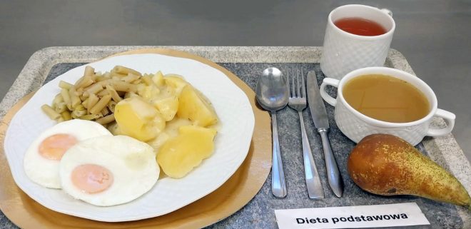 Na tacy stoi talerz z dwoma jajkami sadzonymi, gotowanymi ziemniakami i porcją fasolki szparagowej. Obok znajdują się kubek z herbatą, miseczka z bulionem oraz gruszka. Przy tacy leżą sztućce i karteczka z napisem „Dieta podstawowa”