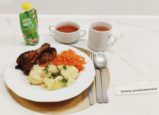 Talerz z obiadem składającym się z dwóch pieczonych pałek kurczaka, gotowanych ziemniaków z koperkiem oraz surówki z marchwi. Obok stoją dwa kubki z napojami i saszetka z musem owocowym. Zestaw sztućców leży po prawej stronie, a przed talerzem znajduje się karteczka z napisem „Dieta podstawowa”