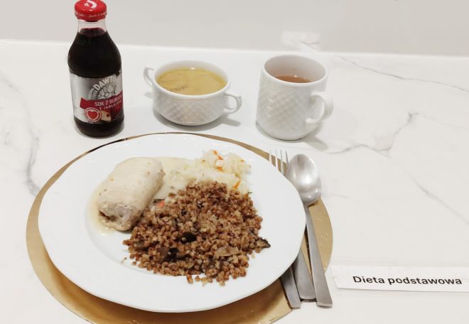 Na białym talerzu ułożonym na złotej podkładce znajduje się porcja kaszy gryczanej z warzywami, gotowany kawałek kurczaka w sosie oraz surówka z białej kapusty. Obok stoją dwie białe filiżanki — jedna z jasną zupą, druga z herbatą — oraz butelka soku z buraka marki Dawtona. Przy talerzu leżą sztućce, a przed nim znajduje się etykieta z napisem Dieta podstawowa
