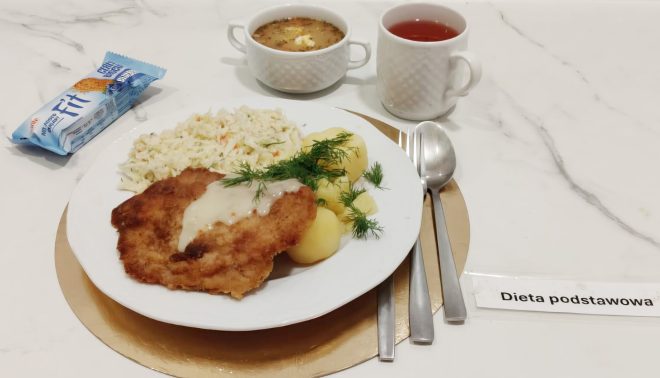 Obiad w diecie podstawowej: na talerzu panierowany kotlet w jasnym sosie, ziemniaki z koperkiem oraz surówka z białej kapusty. Obok znajdują się dwa kubki — jeden z czerwonym kompotem, drugi z zupą jarzynową. Z lewej strony leży batonik typu „Fit”. Zestaw podany jest na złotej podkładce, z widoczną etykietą „Dieta podstawowa”