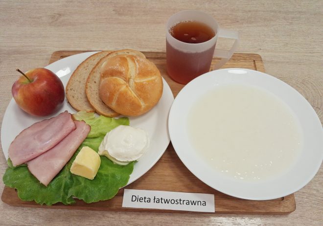 Śniadanie w diecie łatwostrawnej: bułka kajzerka, dwa plasterki chleba pszennego, wędlina, serek twarogowy, masło, liść sałaty, jabłko i herbata zbożowa. Obok talerza znajduje się miseczka z zupą mleczną