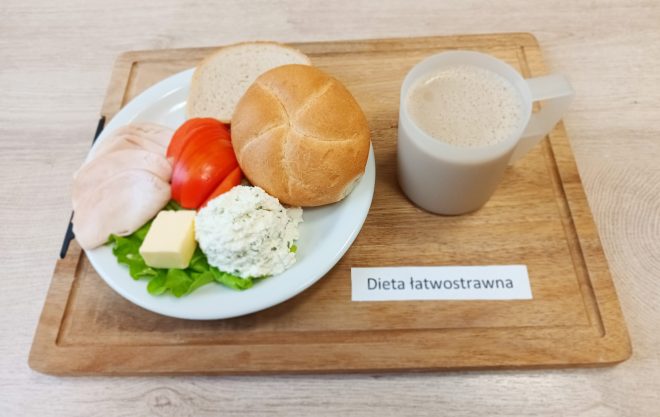 Na tacy położony talerz z jasną bułką i kromką pieczywa, plastrami delikatnej wędliny drobiowej, pomidorem, porcją twarożku z ziołami, kostką masła i listkiem sałaty. Obok przezroczysty kubek z jasnym napojem mlecznym lub kawą z mlekiem. Przed tacą kartka z napisem „Dieta łatwostrawna”