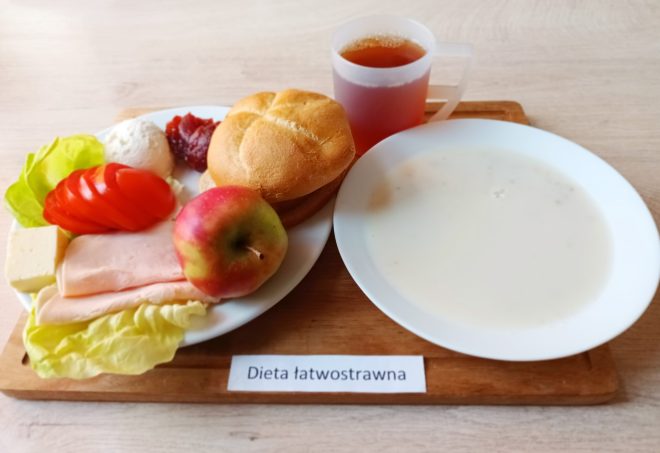 Na tacy śniadanie składające się z bułki, plasterków wędliny drobiowej, sera, pomidora, liścia sałaty, twarożku, porcji dżemu i jabłka. Obok talerz z mlekiem lub zupą mleczną oraz kubek z herbatą. Przed tacką kartka z napisem „Dieta łatwostrawna”