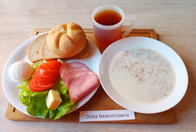 Na tacy z napisem „Dieta łatwostrawna” znajduje się śniadanie: na jednym talerzu podano plasterki pomidora, kilka plastrów chudej wędliny, kulkę białego sera, kostkę masła i liście sałaty, a obok położono kromki jasnego pieczywa i bułkę. Na drugim talerzu widać mleczną zupę z drobną kaszką lub płatkami owsianymi, a obok stoi kubek z herbatą