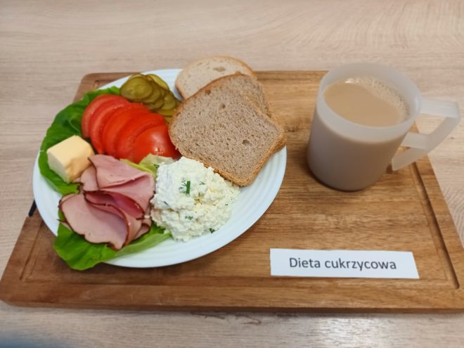 Śniadanie w diecie cukrzycowej: pieczywo, pomidor i ogórek kiszony, plaster sera i masła, plastry wędliny oraz porcja białego twarożku ze szczypiorkiem. Obok kubek kawy z mlekiem.