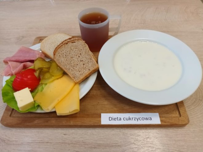 Śniadanie w diecie cukrzycowej: kilka kromek pieczywa, plastry wędliny, ser żółty, plaster masła, ogórek kiszony i pomidor na liściu sałaty. Obok talerz z jasną zupą mleczną oraz kubek herbaty