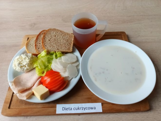 Zestaw śniadaniowy oznaczony jako „Dieta cukrzycowa”. Na talerzu leżą trzy kromki pieczywa razowego, liście sałaty, plasterki pomidora, plastry białej wędliny, twarożek oraz mała kostka masła. Obok znajduje się talerz z jasną zupą mleczną. W tle stoi przezroczysty kubek z ciemnym napojem, najprawdopodobniej herbatą