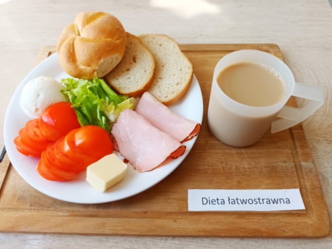 Na białym talerzu podano pieczywo, plasterki pomidora, liść sałaty, plasterki szynki drobiowej, kawałek masła oraz gotowane jajko. Obok znajduje się kubek z jasną kawą z mlekiem. Całość ustawiona na drewnianej tacy z etykietą „Dieta łatwostrawna”