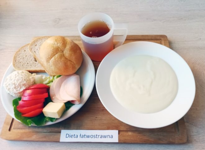 Zestaw śniadaniowy w diecie łatwostrawnej. Na tacy znajduje się talerz z pieczywem — bułką kajzerką i dwoma kromkami chleba — oraz dodatkami: twarożkiem, plastrem szynki drobiowej, kilkoma plasterkami pomidora, masłem i liściem sałaty. Obok stoi kubek herbaty oraz talerz z białą, kremową zupą mleczną lub kleikiem. Przed tacą umieszczono etykietę z napisem ‘Dieta łatwostrawna