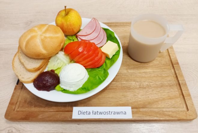 Talerz z pieczywem – bułką i kromkami chleba – oraz dodatkami: plasterkami pomidora, wędliny, kawałkiem masła, twarożkiem, tartą ćwikłą i liśćmi sałaty. Obok kubek z kawą z mlekiem i jabłko. Pod zestawem znajduje się napis ‘Dieta łatwostrawna’