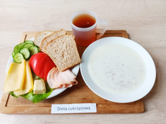 Śniadanie w diecie cukrzycowej: talerz z pieczywem, plastrami sera i wędliny, masłem, warzywami — ogórkiem i pomidorem — oraz listkiem sałaty. Obok stoi kubek herbaty oraz miseczka z białą zupą mleczną z kaszą. Na tacy umieszczono etykietę «Dieta cukrzycowa»