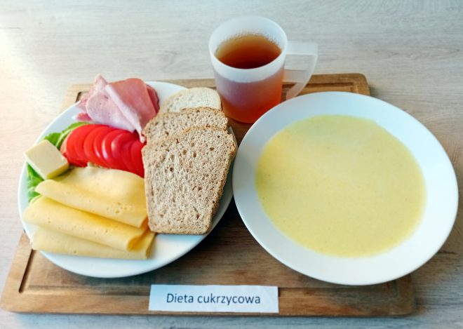 Śniadaniowy zestaw na drewnianej tacy: talerz z pieczywem, plastrami pomidora, sałatą, żółtym serem, masłem i wędliną. Obok znajduje się miska z jasną zupą mleczną oraz kubek z herbatą. Przed tacą leży karteczka z napisem „Dieta cukrzycowa”