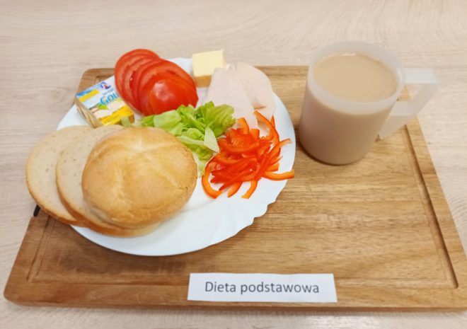 Śniadanie w diecie podstawowej: talerz z bułką i kromkami jasnego pieczywa, masłem, plasterkami pomidora, papryką, sałatą oraz plastrami delikatnej wędliny. Do zestawu dołączony kubek kawy z mlekiem. Posiłek ustawiony na drewnianej tacy z etykietą „Dieta podstawowa”