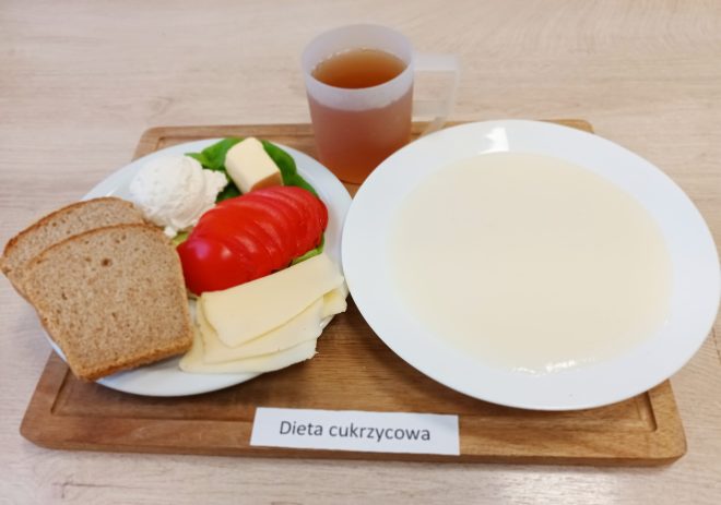 Śniadanie w diecie cukrzycowej: talerz z pieczywem razowym, twarożkiem, plasterkami sera, pomidorem, liściem sałaty i masłem. Obok miseczka z zupą mleczną oraz kubek herbaty. Zestaw znajduje się na drewnianej tacy z etykietą „Dieta cukrzycowa”