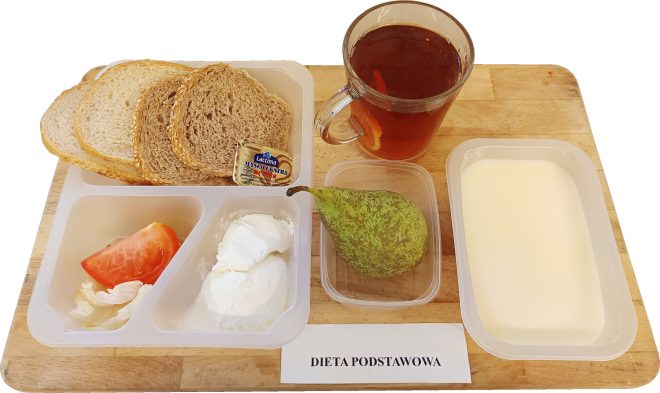 Na drewnianej tacy podany zestaw śniadaniowy. W pojemniku trzy kromki chleba jasnego i ciemnego, porcja masła, plaster pomidora z liściem sałaty oraz serek twarogowy. Obok pojemnik z mlekiem lub napojem mlecznym, szklanka z herbatą z cytryną i pojemniczek z gruszką. Przed tacką kartka z napisem „DIETA PODSTAWOWA”