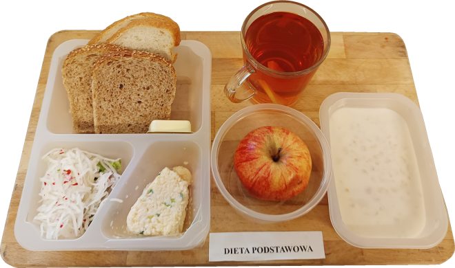 Śniadanie w formie zestawu szpitalnego. Na tacy znajdują się kromki chleba jasnego i ciemnego, kostka masła, porcja pasty twarogowej z ziołami, surówka z białej rzodkwi i marchewki, jabłko, szklanka herbaty z cytryną oraz pojemnik z mlekiem lub kaszką mleczną. Przed tacką karteczka z napisem „DIETA PODSTAWOWA”